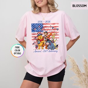 May include: A light pink t-shirt featuring a graphic of the American flag with cartoon characters and the text "1776-2026 America - 250th Anniversary". The shirt has a round neck and short sleeves.