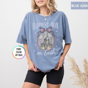 May include: A light blue t-shirt with the text "Happiest Mom on Earth" and a castle graphic. The graphic includes a pink bow, stars, and Mickey Mouse heads. The shirt has short sleeves and a relaxed fit.