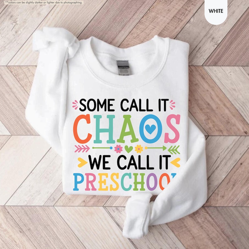 May include: White sweatshirt with the text "SOME CALL IT CHAOS WE CALL IT PRESCHOOL" in colorful letters. The design includes arrows, hearts, and flowers. The sweatshirt is folded on a wooden surface.