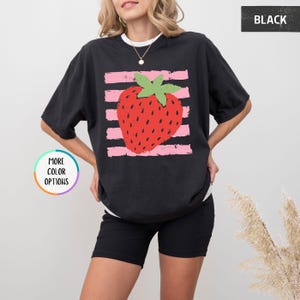 May include: A black short-sleeved t-shirt featuring a graphic of a red strawberry with a green stem centered over horizontal light pink distressed stripes. The shirt has a crew neckline and a relaxed fit. The text BLACK appears in the top right corner.