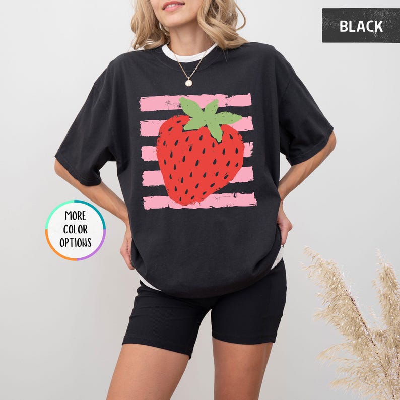May include: A black short-sleeved t-shirt featuring a graphic of a red strawberry with a green stem centered over horizontal light pink distressed stripes. The shirt has a crew neckline and a relaxed fit. The text BLACK appears in the top right corner.