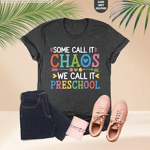 May include: Dark grey heather t-shirt with the text "SOME CALL IT CHAOS WE CALL IT PRESCHOOL" in colorful letters. The image also includes a pair of dark blue jeans, a small pink wallet, and a pair of pink sneakers.