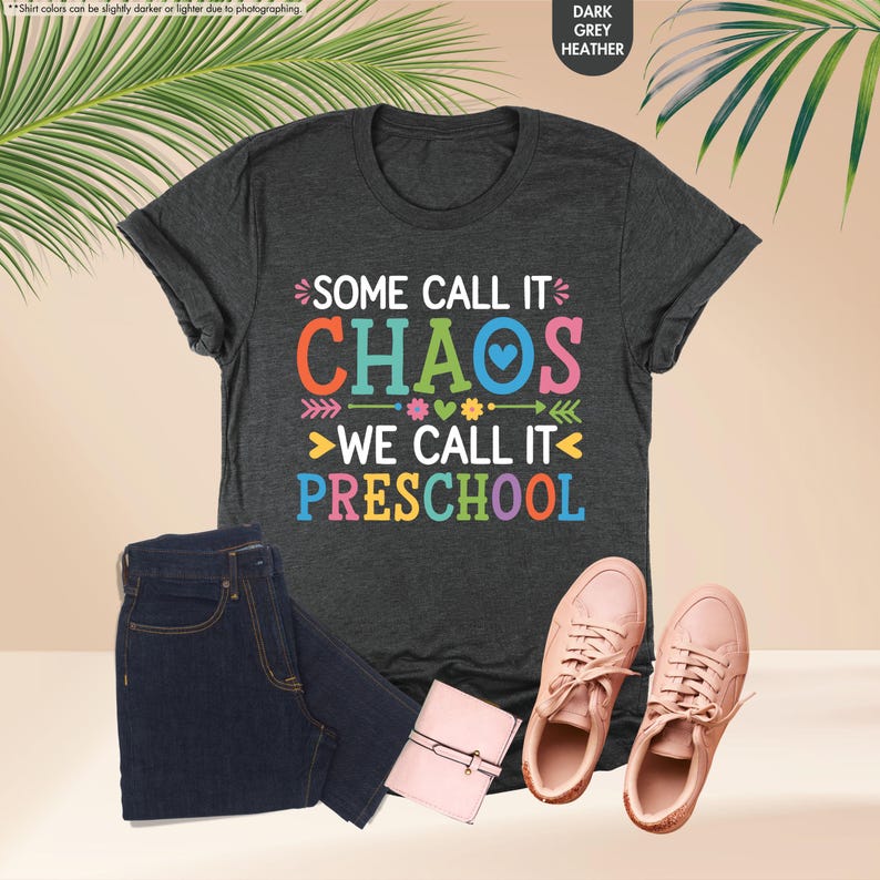 May include: Dark grey heather t-shirt with the text "SOME CALL IT CHAOS WE CALL IT PRESCHOOL" in colorful letters. The image also includes a pair of dark blue jeans, a small pink wallet, and a pair of pink sneakers.