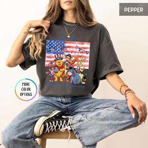 May include: Dark grey t-shirt featuring a graphic of Winnie the Pooh characters in front of an American flag. The flag design incorporates stars and stripes, with the text "1926 97th anniversary" in blue. The shirt is a relaxed fit.