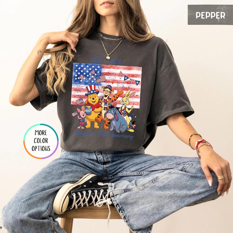 May include: Dark grey t-shirt featuring a graphic of Winnie the Pooh characters in front of an American flag. The flag design incorporates stars and stripes, with the text "1926 97th anniversary" in blue. The shirt is a relaxed fit.