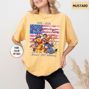 May include: Mustard-colored t-shirt featuring a graphic of Winnie the Pooh and friends in front of an American flag. The shirt commemorates the 250th anniversary of America, with the years 1776-2026 printed above the flag.