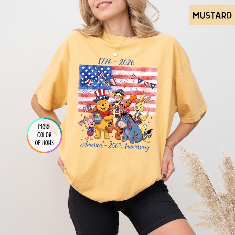 May include: Mustard-colored t-shirt featuring a graphic of Winnie the Pooh and friends in front of an American flag. The shirt commemorates the 250th anniversary of America, with the years 1776-2026 printed above the flag.