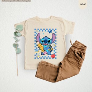May include: A beige t-shirt with a cartoon character holding a pencil, set against a blue and white checkered pattern. The character is blue with large ears and a backpack. Brown trousers are next to the shirt.