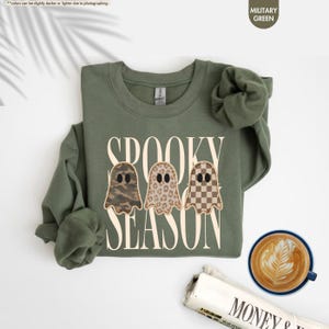 May include: A military green crewneck sweatshirt featuring the text SPOOKY SEASON in large cream letters. Three ghost illustrations are centered, each with a different pattern: camouflage, leopard print, and a brown and cream checkerboard design.