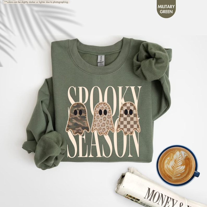 May include: A military green crewneck sweatshirt featuring the text SPOOKY SEASON in large cream letters. Three ghost illustrations are centered, each with a different pattern: camouflage, leopard print, and a brown and cream checkerboard design.