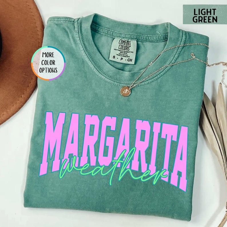 May include: A light green t-shirt with the words "MARGARITA weather" in bright pink and green lettering. The shirt has a crew neck and short sleeves. A gold necklace with a pendant is draped on the shirt.