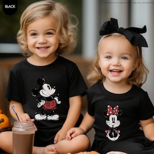 May include: Two children wearing black t-shirts with Disney character graphics. One shirt features Mickey Mouse in red shorts, and the other features Minnie Mouse with a red polka dot bow. Both children are smiling.