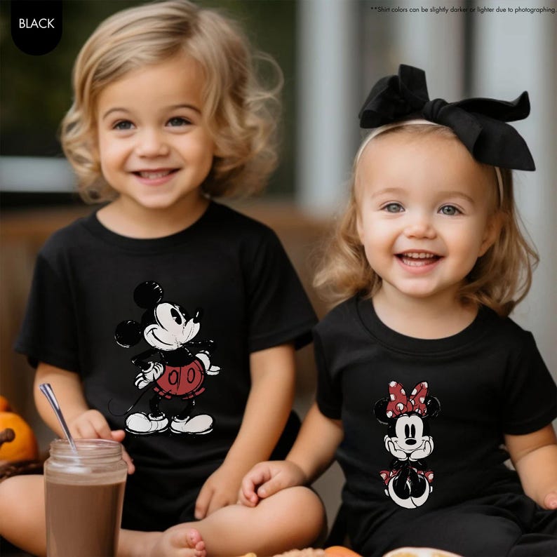 May include: Two children wearing black t-shirts with Disney character graphics. One shirt features Mickey Mouse in red shorts, and the other features Minnie Mouse with a red polka dot bow. Both children are smiling.