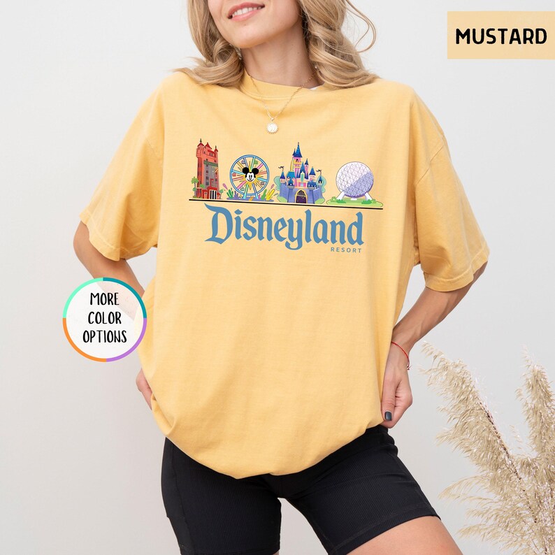 Op de afbeelding: Mosterdkleurig T-shirt met een Disneyland Resort-afbeelding. De afbeelding toont iconische parkafbeeldingen, waaronder een kasteel, een reuzenrad met Mickey Mouse en een bol. De tekst "Disneyland Resort" is blauw gedrukt.