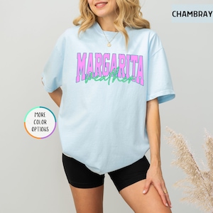 May include: Light blue t-shirt with the text "MARGARITA WEATHER" in pink and green. The shirt is a chambray color and has a round neck. The image also includes a "MORE COLOR OPTIONS" button.
