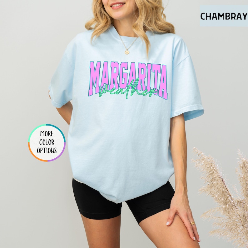 May include: Light blue t-shirt with the text "MARGARITA WEATHER" in pink and green. The shirt is a chambray color and has a round neck. The image also includes a "MORE COLOR OPTIONS" button.