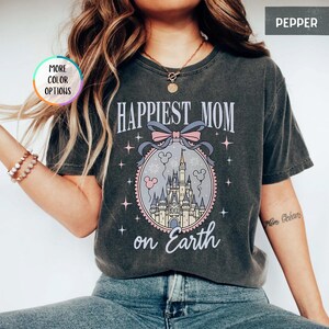 May include: Dark gray t-shirt with the text "HAPPIEST MOM on Earth". The shirt features a graphic of a castle inside an oval frame with a bow and Mickey Mouse icons. The shirt is a Pepper color.