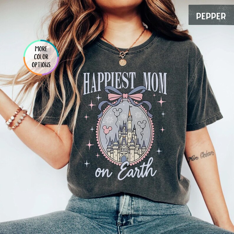 May include: Dark gray t-shirt with the text "HAPPIEST MOM on Earth". The shirt features a graphic of a castle inside an oval frame with a bow and Mickey Mouse icons. The shirt is a Pepper color.