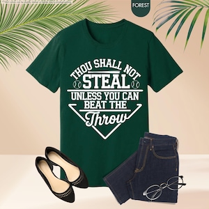 May include: A forest green t-shirt with the text "THOU SHALL NOT STEAL UNLESS YOU CAN BEAT THE THROW" in white. The shirt is paired with dark wash jeans, black flats, and eyeglasses. The shirt has a baseball theme.