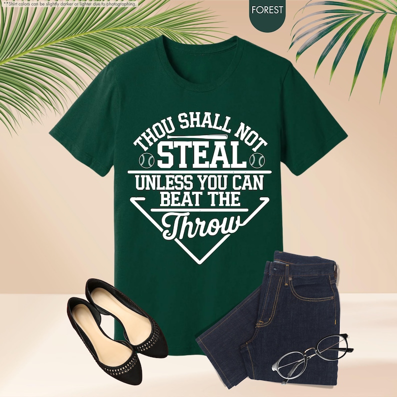 May include: A forest green t-shirt with the text "THOU SHALL NOT STEAL UNLESS YOU CAN BEAT THE THROW" in white. The shirt is paired with dark wash jeans, black flats, and eyeglasses. The shirt has a baseball theme.