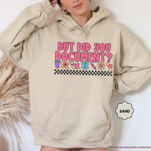 Puede incluir: Sudadera color arena con el texto "BUT DID YOU DOCUMENT?" en letras rosas y naranjas. Debajo del texto hay gr&aacute;ficos coloridos con tem&aacute;tica m&eacute;dica y un borde a cuadros blanco y negro.