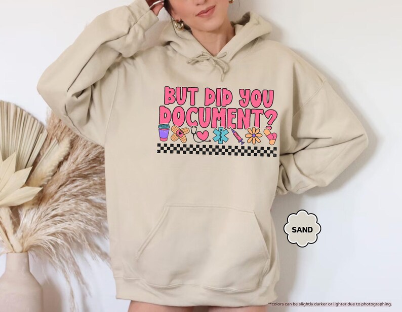 Puede incluir: Sudadera color arena con el texto "BUT DID YOU DOCUMENT?" en letras rosas y naranjas. Debajo del texto hay gr&aacute;ficos coloridos con tem&aacute;tica m&eacute;dica y un borde a cuadros blanco y negro.
