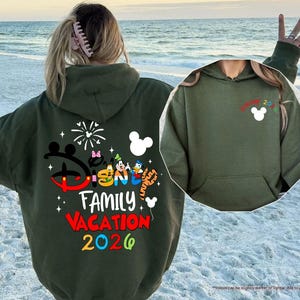 Puede incluir: Una sudadera con capucha de color verde militar que presenta un gr&aacute;fico grande en la espalda con el texto Disney Family Vacation 2026, personajes de dibujos animados y fuegos artificiales. El pecho muestra un icono de Mickey Mouse y Disney 2026.