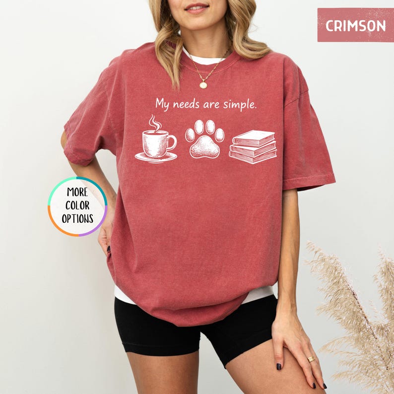 May include: Crimson-colored t-shirt with the text "My needs are simple." and illustrations of a coffee cup, paw print, and books. The shirt has short sleeves and a relaxed fit. "More color options" is also visible.