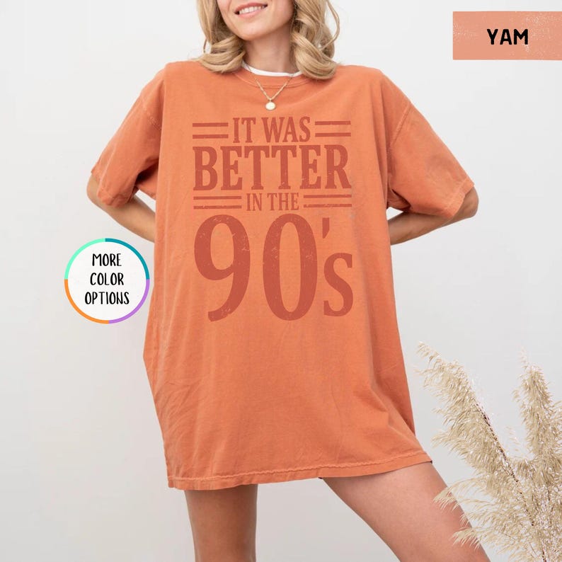 May include: An oversized burnt orange t-shirt featuring the dark orange text "IT WAS BETTER IN THE 90's" printed on the front. The shirt has short sleeves and a crew neckline. A circular graphic on the left side displays the text "MORE COLOR OPTIONS".