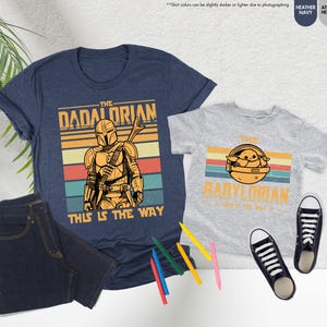 Puede incluir: Dos camisetas, una azul marino y otra gris jaspeado, con gr&aacute;ficos a juego. La camiseta azul marino dice "The Dadalorian" y "This is the Way". La camiseta gris dice "The Babylorian" y "This is the Way". Tambi&eacute;n se ven unos vaqueros oscuros y zapatillas negras.