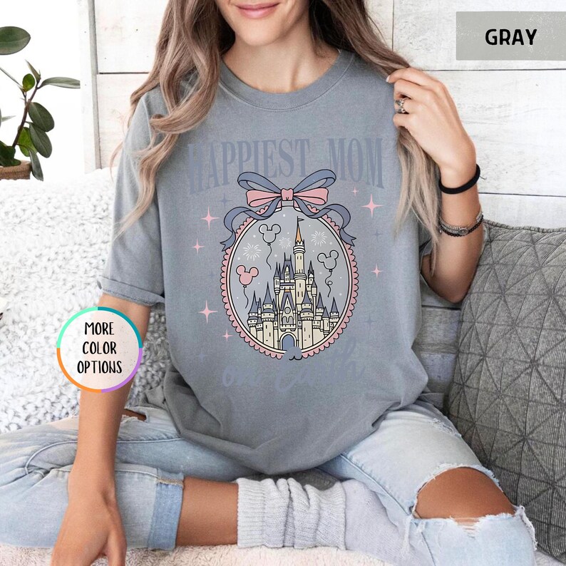 May include: Gray t-shirt with the text "Happiest Mom" above a castle design inside a pink and blue frame. The design includes a bow, stars, and Mickey Mouse balloons. The shirt is a comfortable, casual style.