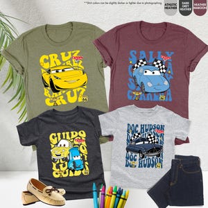 May include: Four short-sleeved t-shirts in olive green, maroon, dark grey, and heather grey, each with a cartoon car design. The shirts feature the names "Cruz", "Sally", "Guido", and "Doc Hudson" and the Route 66 logo.
