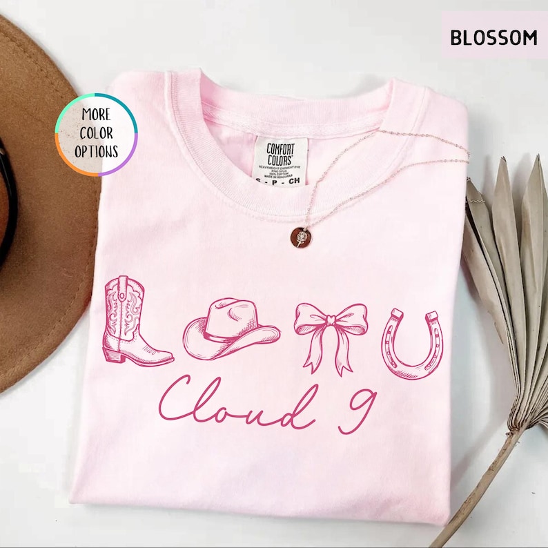 May include: A light pink t-shirt with a crew neck, featuring a cowboy boot, hat, bow, and horseshoe design, with the words "Cloud 9" in a cursive font. A gold necklace with a pendant is draped across the shirt.