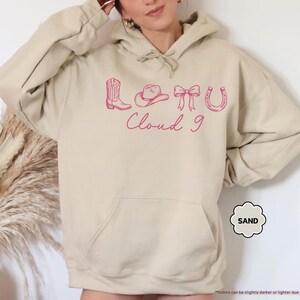 May include: A beige hoodie with a front pocket and a hood with a drawstring. It has a pink graphic of a cowboy boot, hat, bow, horseshoe, and the words "Cloud 9". The hoodie is a relaxed fit.