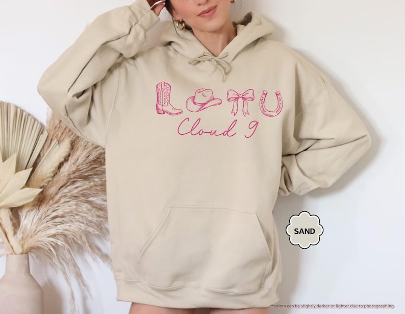 May include: A beige hoodie with a front pocket and a hood with a drawstring. It has a pink graphic of a cowboy boot, hat, bow, horseshoe, and the words "Cloud 9". The hoodie is a relaxed fit.