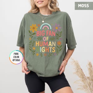 May include: Moss green t-shirt with the text "BIG FAN OF HUMAN RIGHTS" in colorful letters and a rainbow graphic. The shirt is adorned with floral and leaf designs. The shirt is a comfortable, casual style.