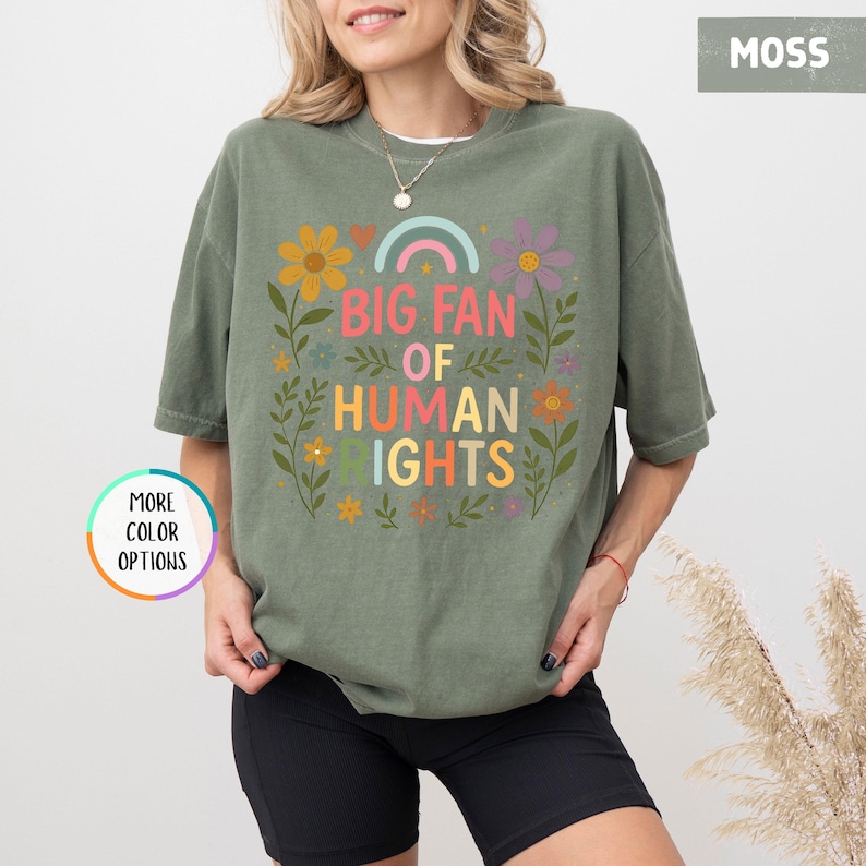 May include: Moss green t-shirt with the text "BIG FAN OF HUMAN RIGHTS" in colorful letters and a rainbow graphic. The shirt is adorned with floral and leaf designs. The shirt is a comfortable, casual style.