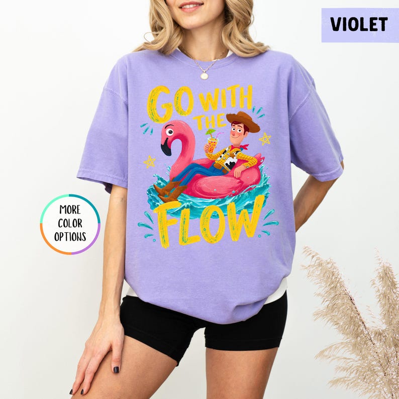 Puede incluir: Una camiseta violeta de manga corta con un gr&aacute;fico de un vaquero sentado en un flotador de flamenco rosa sobre agua azul. El texto "GO WITH THE FLOW" est&aacute; impreso en amarillo sobre y bajo el personaje. Un c&iacute;rculo dice "MORE COLOR OPTIONS" y un recuadro "VIOLET".