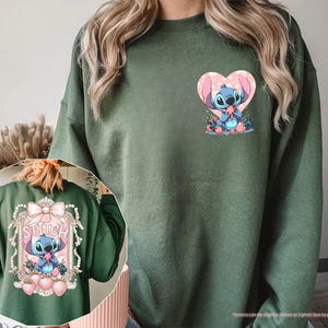 Puede incluir: Sudadera verde militar con un dise&ntilde;o de Stitch. La parte delantera muestra a Stitch dentro de un coraz&oacute;n rosa, mientras que la parte trasera presenta a Stitch dentro de un marco decorativo con la palabra "Stitch". El dise&ntilde;o incluye fresas rosas y flores azules.