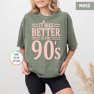 May include: A moss green short-sleeve t-shirt featuring the light pink text IT WAS BETTER IN THE 90's printed on the front. The shirt has a crew neckline and a relaxed fit. A circular graphic with the text MORE COLOR OPTIONS is visible.