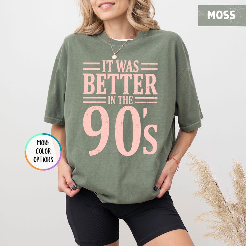 May include: A moss green short-sleeve t-shirt featuring the light pink text IT WAS BETTER IN THE 90's printed on the front. The shirt has a crew neckline and a relaxed fit. A circular graphic with the text MORE COLOR OPTIONS is visible.