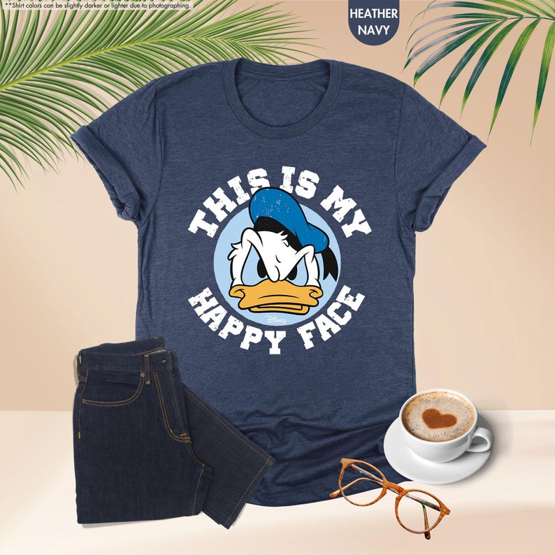 Op de afbeelding: Marineblauw T-shirt met de tekst "THIS IS MY HAPPY FACE" en een afbeelding van Donald Duck. De afbeelding toont ook een donkerblauwe jeans, een koffiekopje met een hart en een bril met schildpadmotief.