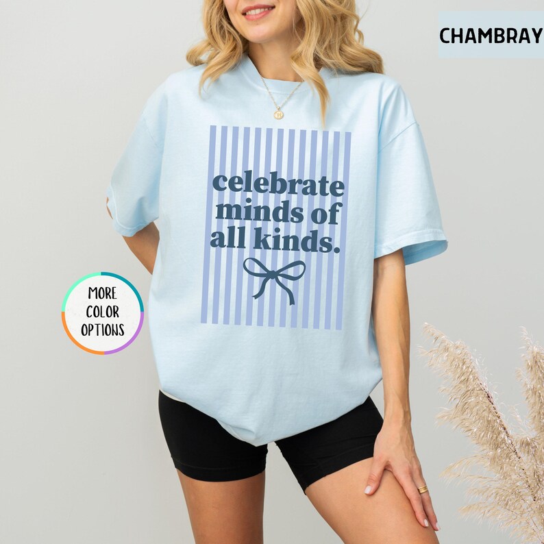 Puede incluir: Camiseta azul claro con el texto "celebrate minds of all kinds" en un rect&aacute;ngulo azul con rayas verticales y un lazo. La camiseta es de color chambray y tiene "more color options" en un c&iacute;rculo.