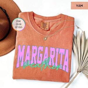 May include: A yam-colored Comfort Colors t-shirt with the words "MARGARITA weather" in pink and teal lettering. A gold necklace with a pendant is draped on the shirt. A brown hat and a dried palm leaf are in the background.