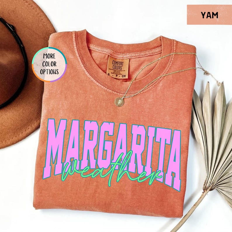 May include: A yam-colored Comfort Colors t-shirt with the words "MARGARITA weather" in pink and teal lettering. A gold necklace with a pendant is draped on the shirt. A brown hat and a dried palm leaf are in the background.