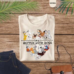 May include: A cream-colored t-shirt featuring a heart-shaped arrangement of cartoon dogs. The shirt has the text "Life is Better With Dogs" in black. The shirt is displayed on a wooden surface with a pair of blue jeans and a brown purse.