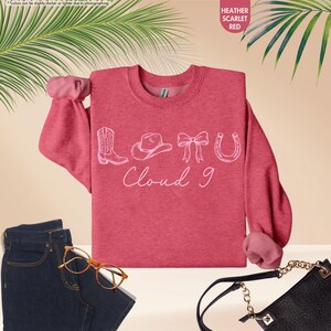 May include: Heather scarlet red sweatshirt featuring a white graphic of a cowboy boot, hat, bow, and horseshoe, with the text "Cloud 9" underneath. The image includes blue jeans, glasses, and a black handbag.