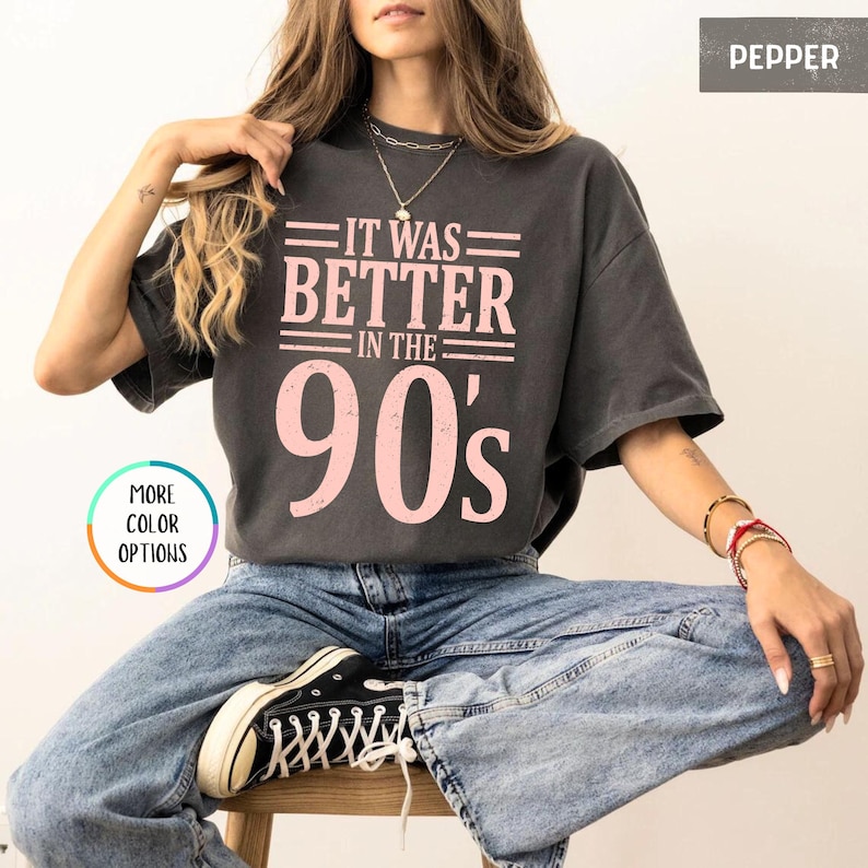 May include: A charcoal gray short-sleeve t-shirt featuring the light pink text IT WAS BETTER IN THE 90's printed on the front. The shirt has a crew neckline and a relaxed fit, paired with blue denim jeans and black and white canvas sneakers.