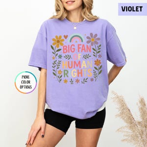 May include: A violet-colored t-shirt with the text "BIG FAN OF HUMAN RIGHTS" in colorful letters, surrounded by floral and rainbow graphics. The shirt has short sleeves and a relaxed fit. Additional text on the shirt says "MORE COLOR OPTIONS".