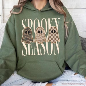 May include: A military green hooded sweatshirt featuring the text SPOOKY SEASON in large cream letters. Centered between the words are three ghost illustrations with black oval eyes, each featuring a different pattern: camouflage, leopard print, and a brown checkerboard.